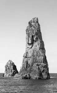 A black and white image captures a serene ocean scene with two towering rock formations, one resembling a human face and the other a faceless rock, surrounded by calm water and a clear sky, evoking a sense of solitude and tranquility.